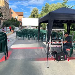 aufgebautes Open Air Kino, eine große weiße Leinwand, davor Kunststoffstühle in Reihen und ein Pavillon unter dem die Technik steht