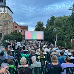 besetzte Stuhlreihen vor einer Leinwand auf der ein Film läuft im Freien
