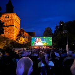 Foto auf dem die Stimmung beim Open Air Konzert eingefangen wurde. Es ist dunkel, Menschen sind schemenhaft zu erkennen wie sie auf den Stühlen vor einer großen Leinwand sitzen und einen Film schauen, links neben der Leinwand ist ein beleuchteter Wachturm zu sehen.
