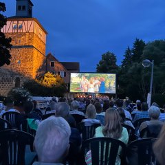 Foto auf dem die Stimmung beim Open Air Konzert eingefangen wurde. Es ist dunkel, Menschen sind schemenhaft zu erkennen wie sie auf den Stühlen vor einer großen Leinwand sitzen und einen Film schauen, links neben der Leinwand ist ein beleuchteter Wachturm zu sehen.