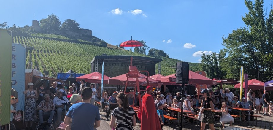 Eine Gruppe von Menschen auf einem Fest. Es sind Bänke, Schirme und ein roter Stuhl zu sehen. Im Hintergrund sieht man eine Bühne und die Burg Weibertreu