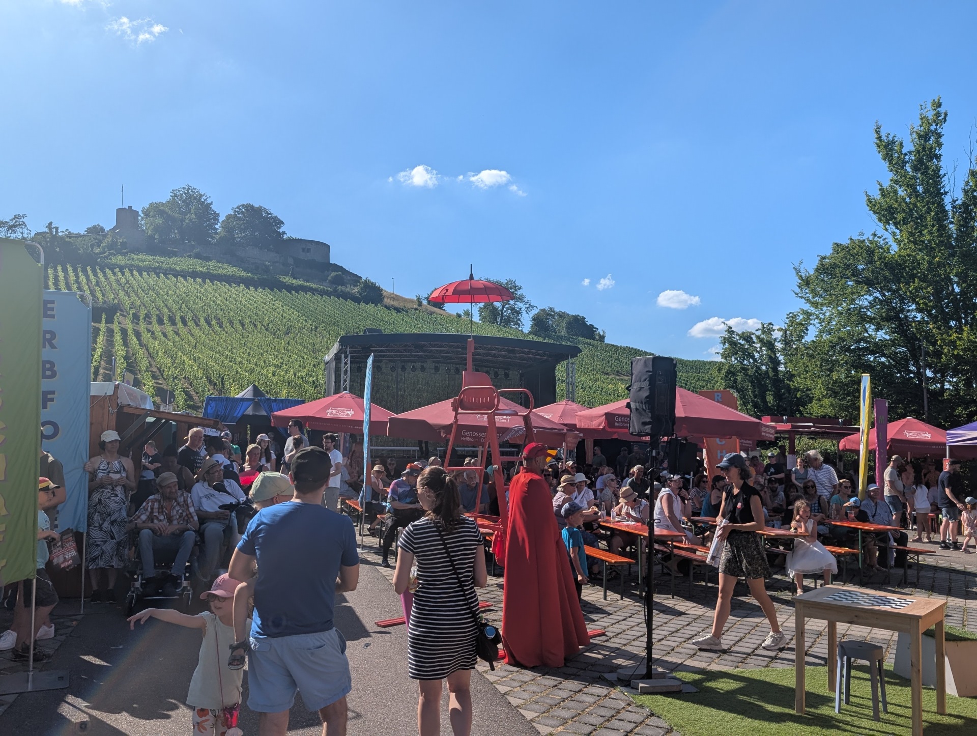 Eine Gruppe von Menschen auf einem Fest. Es sind Bänke, Schirme und ein roter Stuhl zu sehen. Im Hintergrund sieht man eine Bühne und die Burg Weibertreu