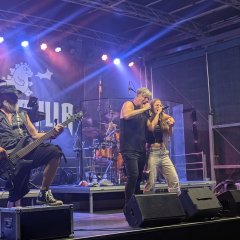 Auf dem Bild ist eine Bühne mit einer Rockband zu sehen: Eine Sängerin und ein Sänger, ein Mann, der Keyboard spielt und zwei Männer, die Gitarre spielen
