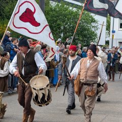 Bauernmarsch auf den Lindenplatz; Eine Gruppe von Personen marschiert in einer Art langem Aufmarsch auf einer Straße. Im Hintergrund des Geschehens stehen Schaulustige und beobachten das Spektakel. Im Vordergrund des Bildes marschieren Bauern, gekleidet in einfachen zeitgenössischen Kostümen, auf den Lindenplatz zu. V.L.n.R. Ein Jugendlicher, über die Schulter gestützt trägt er eine dreieckige Fahne mit einem Bundschuh drauf, dass Zeichen der aufständischen Bauern von 1525. Neben ihm läuft ein Mann, um die Hüfte gebunden eine Trommel, in den beiden Händen die Schläger, mit seinem Trommeln kündigt er den Zug an. Neben diesem läuft ein weiterer etwas älterer Mann, er stell Jäcklein Rohrbach dar, den Anführer der Aufständischen. In der Hand trägt er eine Lanze. In zweiter Reihe v.L.n.R. Ein weiterer Bauer folgt mit einer weiteren Fahne des Bundschuhes über der Schulter gestützt. Neben diesem läuft ein Mann im Priestergewand mit einem Ferula (Kreuzstab) zum Himmel gestreckt. Einige Meter hinter dieser ersten Gruppe folgt eine weitere, größere Gruppe Bauern. In den Händen haben Sie Lanzen und Speere. In Ihrer Mitte führen Sie einen Gefangenen. Ihm sind die Hände mit einem Seil gebunden, einer der Bauern führt Ihn an diesem Seil die Straße entlang. Der Gefangene sticht durch seine rote und sehr aufwändige Kleidung hervor, er stellt den gefangenen Grafen Helfenstein dar.
