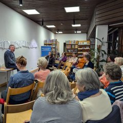 Vortrag von Herrn Dr. Liebig Das Bild ist in der Stadtbücherei Weinsberg aufgenommen. Im rechten Teil des Bildes steht ein Tisch an einer Wand. Auf diesem Tisch sitzt Herr Dr. Liebig, der mit und zu seinen Zuhörerinnen spricht. In einem Halbkreis um den Tisch auf der linken verteilt sind Stühle aufgestellt. Auf diesen Stühlen sitzen einige Frauen und hören dem Vortrag zu. Im Hintergrund des Bildes stehen einige volle Bücherregale, welche mit der Wand auf der rechten Seite des Bildes abschließen. Die Wand geht in eine Glasfront über.