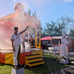 Auftritt der Roadshow Das Bild ist auf dem Parkplatz am grasigen Hag aufgenommen, es zeigt eine Kunstaufführung im Rahmen der Roadshow. Der vordere linke Bereich des Bilds ist mit einem Teppich aus grünem Kunstgras ausgefüllt. An der vorderen linken und vorderen rechten Ecke stehen jeweils Podeste. Auf diesen Podesten stehen Personen. Links ein Mann, rechts eine Frau. Beide tragen weiße Kleidung. Sie strecken beide den innen liegenden Arm zu einer Faust nach oben. Ihre Gesichter sind Ernst. Hinter den beiden, das ganze linke Bild im Hintergrund ausfüllend steht ein sechseckiger Pavillon. Er ist in abwechselnd gelb und orange gestrichen, ebenso ist das Stoffdach. Auf dem Dacht thront eine große goldene Kugel im Zentrum. Auf der Bühne selber steht eine Person in dunklen Klamotten. Von der hinteren Seite bläst eine Person im schwarzen Umhang Nebel in den Pavillon. Der Nebel ist Rot. Hinter dem Pavillon steht, bis zum Ende des Bildes auf der rechten Seite eine Reihe von Hütten und Zelten zum Verkauf von Speisen. Vor dem letzten Stand steht ein XXL-Tischkicker. Rechts im Vordergrund des Bildes sitzen und stehen Kinder und schauen der Aufführung zu.