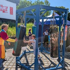 Spiel bei der Roadshow Das Zentrum des Bildes ist ein blaues, hölzernes, hohes Sechseck. An einem Pfeiler hängt eine große rote „2“ und eine Erklärung der Mitmach-Station Zwischen den Ecken hängen an Seilen Boxsäcke. Die Boxsäcke haben verschiedene Muster auf sich abgebildet. Bauerngruppe oder einzelne Bauern in verschiedenen Darstellungen. In der Mitte steht ein kleines Mädchen und drückt gegen einen der Säcke. Ihr gegenüber im linken Bereich des Bildes steht ein Mann. Er drückt wiederum gegen den Sack. Der Mann trägt ein buntes Gauklerkostüm mit langen blauen Socken. Von links bis in die Mitte, hinter das Sechseck erstreckt sich eine Mauer, sie stützt einen kleinen Hang mit einer Grasfläche ab. Auf dem Hang steht ein weißer LKW. Auf seiner Seite ist „UFFRUR! …on the road“ in roter Schrift zu lesen. Hinter ich ist das Kirchenschiff der Johanneskirche hinter einem Baum zu erkennen. Weitere Bäume säumen den Hintergrund bis schließlich auf der oberen rechten Seite Weinberge zu sehen sind. Rechts im Bild, bis in die Mitte stehen, hinter dem Sechseck ein Pavillon und eine Holzhütte mit weiteren Mitmach-Stationen. Dort stehen drei Personen sie dem kleinen Mädchen bei seiner Station zuschauen.