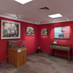 Weibertreumuseum Zu sehen ist ein Raum im Weibertreumuseum in Weinsberg. Der Raum hat eine rote Wandfarbe. An der Wand stehen zwei Vitrinen mit Ausstellungsstücken. An der Wand hängen drei Bilder und eine Infotafel.