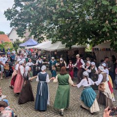 Bauerntanz Unter einem großen Kastanienbaum auf dem Ökolampadiusplatz an der Johanneskirche, im Zentrum des Bildes, hat eine Gruppe Personen einen Kreis gebildet, indem sie sich Hand in Hand halten. Die Personen sind altersmäßig bunt gemischt und in mittelalterlich-bäuerlichen Gewändern gekleidet. Gemeinsam führen sie einen zeitgenössischen Tanz durch vor und zurücklaufen auf. Hinter ihnen stehen Musiker mit Blas- und Streichinstrumenten und begleiten den Tanz musikalisch. Am Rande des Platzes sind Stände aufgebaut, hier verkaufen Händler ihre Waren auf dem Handwerkermarkt. Um die Tänzer herum stehen Schaulustige und schauen dem Tanz zu.