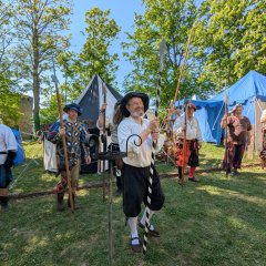 Lager auf der Weibertreu Im linken Hintergrund des Bildes sehen wir den ehemaligen Treppenturm auf der Brug Weibertreu. Wir befinden uns auf dieser. Vor einer Baumreihe vor dem Turm ist auf der Burg ist ein mittelalterliches Lager aufgebaut. Über das Bild verteilt sind verschiedene Zelte aus Stoff aufgebaut. Im Vordergrund steht eine Reihe Männer. Sie tragen mittelalterliche Gewänder und Hauben. In ihren Händen halten sie, auf den Boden abgestützt, Hellebarden. Ganz vorne im Bild steht ein älterer Mann mit einem langen Speer und in den beiden Händen und erklärt über das historische Lager und die Bewaffnung.