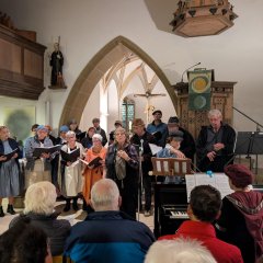 Gellmersbach im Bauernkrieg Auf dem Bild zu sehen ist eine Gruppe Sängerinnen und Sänger, die bäuerlich gekleidet sind, in der Leonhardskirche in Gellmersbach. Im Vordergrund ist das sitzende Publikum zu sehen. Auf der rechten Seite spielt eine Frau am Klavier.