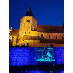 Beleuchtete Kirche Zu sehen sind der Glockenturm, sowie das ihn umgebende Kirchenschiff der Johanneskirche in der späten Abenddämmerung, der Himmel ist nur noch dunkelblau. Die Kirche ist im oberen Zentrum des Bildes. Sie ist mit Scheinwerfern hell erleuchtet. Im unteren Teil des Bildes steht die alte Stadtmauer. Sie ist in dunkelblauer Farbe angestrahlt. Auf sie wird eine Projektion geworfen. In der unteren Rechten Ecke des Bildes ist darauf in weißer Farbe das Logo der Stadtkappelle Weinsberg so wie das Logo des Erinnerungsjahres 2025 zu sehen.
