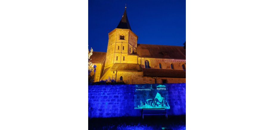 Beleuchtete Kirche Zu sehen sind der Glockenturm, sowie das ihn umgebende Kirchenschiff der Johanneskirche in der späten Abenddämmerung, der Himmel ist nur noch dunkelblau. Die Kirche ist im oberen Zentrum des Bildes. Sie ist mit Scheinwerfern hell erleuchtet. Im unteren Teil des Bildes steht die alte Stadtmauer. Sie ist in dunkelblauer Farbe angestrahlt. Auf sie wird eine Projektion geworfen. In der unteren Rechten Ecke des Bildes ist darauf in weißer Farbe das Logo der Stadtkappelle Weinsberg so wie das Logo des Erinnerungsjahres 2025 zu sehen.