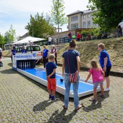 Das Bild zeigt eine Veranstaltung im Freien an einem sonnigen Tag. Im Mittelpunkt steht eine mobile Kegelbahn mit der Aufschrift „TSV 1866 Weinsberg Kegeln“. Mehrere Menschen – darunter Erwachsene und Kinder – schauen zu oder nehmen am Kegelspiel teil. Weitere Details auf dem Bild: • Die Kegelbahn ist blau-weiß gestaltet und steht auf einem gepflasterten Platz. • Vor der Bahn steht ein Junge, daneben eine Frau und ein kleines Mädchen in pinker Kleidung. • Im Hintergrund sind Vereinsmitglieder und Helfer in blauen Shirts zu sehen. • Rechts oben stehen mehrere Fahrräder, einige Personen tragen Fahrradhelme. • Es gibt Bäume, Sonnenschirme und Gebäude im Hintergrund. • Das Wetter wirkt warm und freundlich mit blauem Himmel und Sonnenschein.