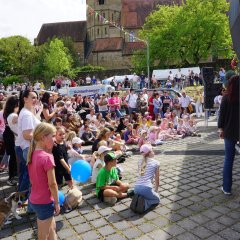 Das Bild zeigt eine große Menschenmenge bei einer öffentlichen Veranstaltung im Freien an einem sonnigen Tag. Viele Erwachsene und Kinder sitzen oder stehen auf einem gepflasterten Platz und schauen in Richtung einer Bühne oder eines Auftrittsbereichs auf der rechten Seite des Bildes. Dort sind große schwarze Lautsprecher aufgebaut. Im Vordergrund sitzen zahlreiche Kinder auf dem Boden, einige halten blaue Luftballons. Links unten liegt außerdem ein kleiner Hund neben den Zuschauern. Die Stimmung wirkt fröhlich und entspannt, viele Menschen lachen oder beobachten aufmerksam das Geschehen. Im Hintergrund sind große grüne Bäume sowie ein historisches Gebäude mit roten Ziegeldächern zu sehen, vermutlich eine Kirche oder ein Kloster. Über dem Platz hängen bunte Wimpelketten, was auf ein Fest oder Stadtfest hinweist. Insgesamt vermittelt das Bild den Eindruck eines Familien- oder Kulturfestes mit Live-Unterhaltung für Kinder und Erwachsene.