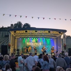 Eine Band namens ROCKZYLLA spielt auf einer Bühne bei Sonnenuntergang vor einem Publikum. Hinter der Bühne erstrecken sich Weinberge auf einem Hügel mit der Burg Weibertreu auf dem Gipfel. Girlanden mit bunten Fahnen hängen über der Veranstaltung.
