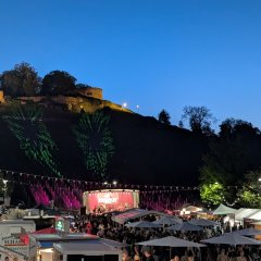 Das Bild zeigt ein Outdoor-Fest in der Dämmerung, bei dem sich eine Menschenmenge vor der Bühne versammelt hat. Der Himmel ist tiefblau. Die Bühne ist mit festlichen Lichterketten geschmückt, die zur Atmosphäre eines Festes beitragen. Im Hintergrund auf einem Hügel die Burg Weibertreu. Die Gesamtstimmung der Szene ist fröhlich und gesellig und deutet auf einen Moment hin, in dem Menschen zusammenkommen, um Musik, Kultur und die Gesellschaft des anderen unter offenem Himmel zu genießen.