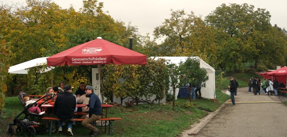 An einer Weinwanderstation sitzen mehrere Gäste an Biertischgarnituren unter roten und weißen Sonnenschirmen. Im Hintergrund stehen weitere Verkaufsstände und Zelte entlang eines befestigten Weges. Die umliegenden Bäume und Weinreben zeigen herbstlich gelb-grüne Blätter. Der Himmel ist bedeckt.