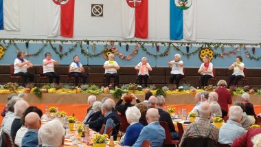 8 Frauen mit bunten Tüchern sitzen auf Stühlen und bewegen die Arme zu Klängen des „Cancan“