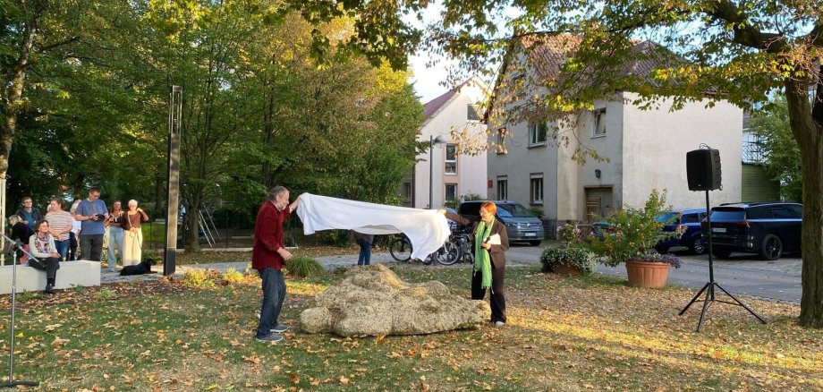Mittig im Bild liegt eine Steinskulptur auf einer Rasenfläche. Links davon steht Karl-Heinrich Lumpp, rechts davon Bürgermeisterin Birgit Hannemann. Die beiden ziehen ein weißes Tuch von der Skulptur. Links im Hintergrund stehen Menschen, die zu den beiden schauen.